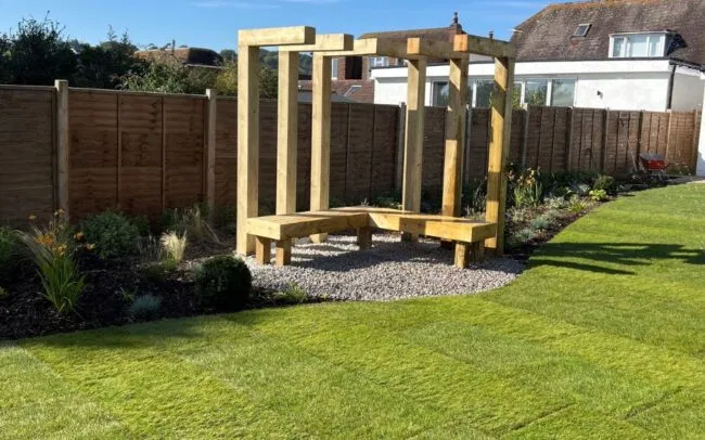 A fire pit area with gravel and wooden bench and structure for climbing plants in a garden in Findon Valley